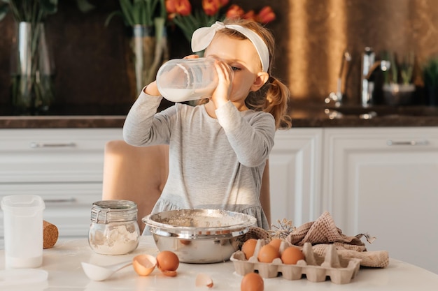 Feliz niña emocionada horneando pastel para Pascua Niño emotivo cocinar postre presente para el día de la madre en la cocina blanca y soleada Panadería casera Botella de leche huevos harina sobre la mesa beber leche