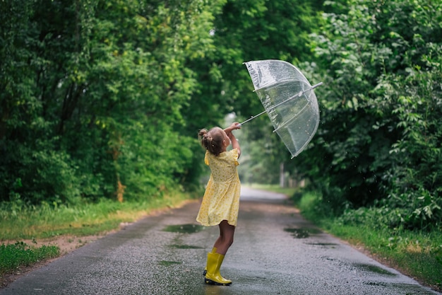 Feliz niña emocional con paraguas transparente en vestido amarillo y botas de lluvia en el verano del parque