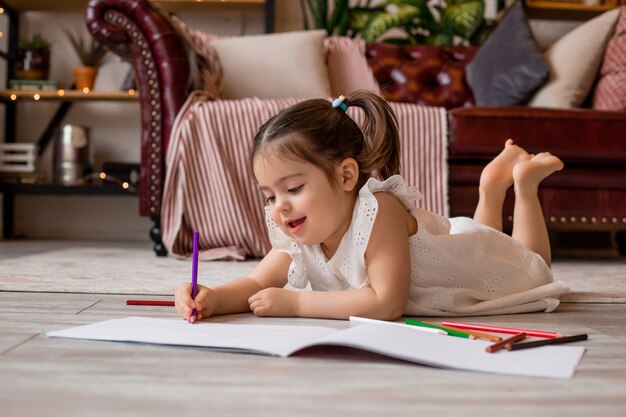 Feliz niña se encuentra en el piso de la casa y dibuja con lápices de colores. desarrollo del niño Quédate en casa