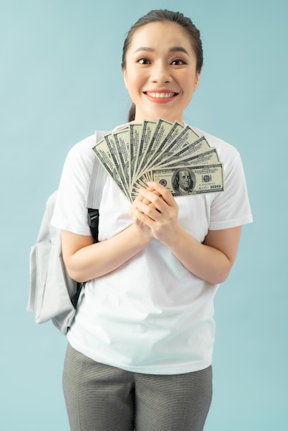 Feliz niña estudiante aislada sobre fondo azul, mantenga ventilador de dinero en efectivo en billetes de dólar haciendo gesto ganador