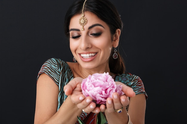 Feliz niña hindú con maquillaje vistiendo vestido tradicional indio sari y joyería étnica sosteniendo flor aislada sobre pared negra