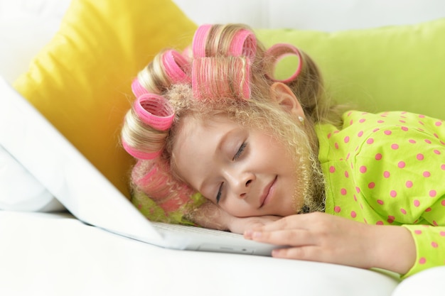 Feliz niña linda en rizadores de pelo durmiendo en su cama con portátil