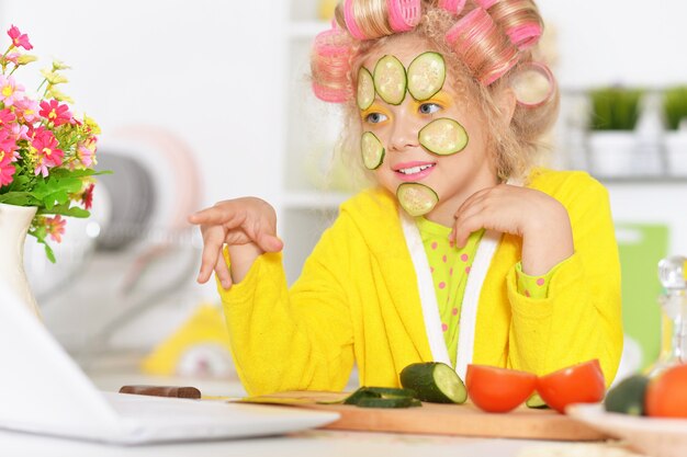 Feliz niña linda en rizadores de pelo con ordenador portátil y verduras