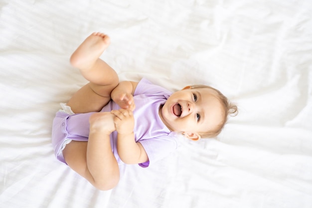 Feliz niña linda en un traje se encuentra de espaldas en una cama sobre sábanas blancas, sonríe, juega con sus piernas. El concepto de productos para bebés.