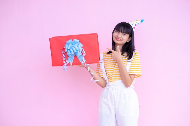 Feliz niña mantenga sombrero de fiesta de desgaste de regalo de caja roja sobre fondo rosa