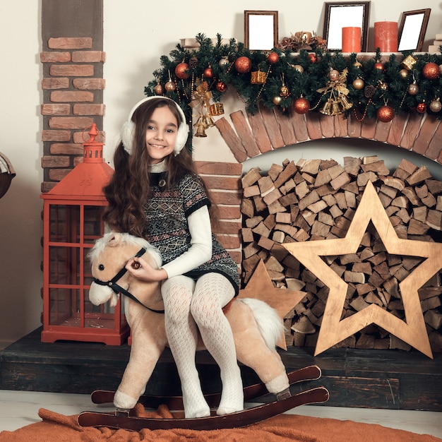 Feliz niña morena con el pelo largo sentado en un caballo de juguete en Navidad decorado habitación.