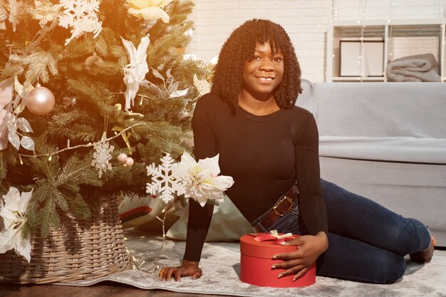 Feliz niña negra con pelo rizado se sienta cerca de un árbol de Navidad y tiene un regalo, mirando a la cámara