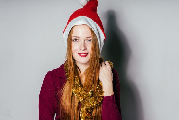 Feliz niña pelirroja con una gorra roja como Santa Claus y con un oropel dorado en el cuello esperando la Navidad