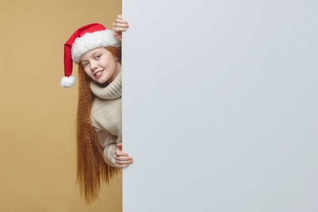 Feliz niña pelirroja con un gorro de Papá Noel con una cartelera blanca