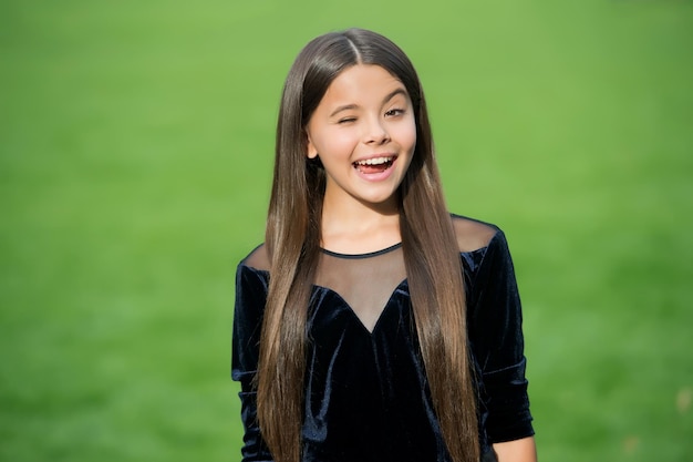 Feliz niña pequeña con el pelo largo en la moda desgaste sonrisa con guiño cara hierba verde verano soleado al aire libre odontología