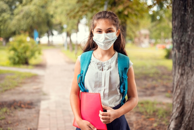 Feliz niña de pie en el parque con mochila a la espalda, con máscara médica