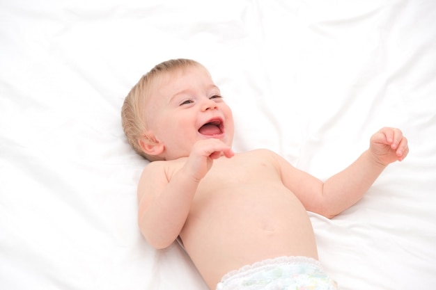 Feliz niña rubia caucásica de aproximadamente 1 año de edad riendo acostado boca arriba sobre ropa de cama blanca. Niño divirtiéndose antes de ir a la cama o después de despertarse. Adorable lindo niño sonriente en sábanas de luz.