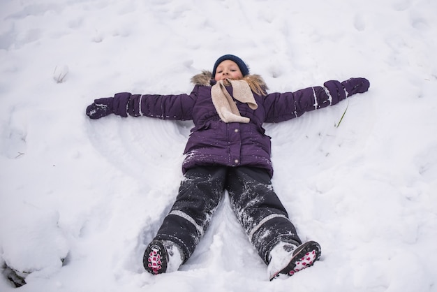 Feliz niña rubia se encuentra en la nieve y hace un ángel, niño divirtiéndose jugando con nieve en día de invierno