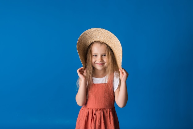 Feliz niña rubia con un sombrero de paja y un vestido de verano se encuentra sobre una superficie azul con una copia del espacio