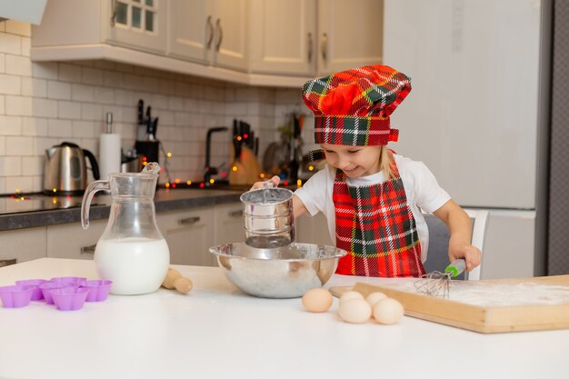 Feliz niña rubia sonriente en un traje de chef amasa la masa para hornear en la cocina de Navidad