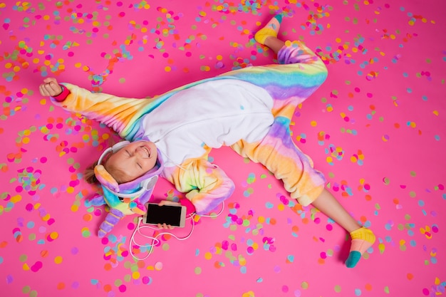 Feliz niña rubia en unicornio kigurumi escucha música con smartphone en la mano sobre una superficie rosa