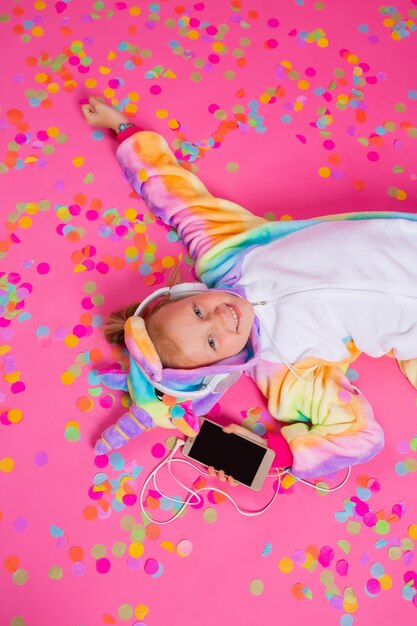 Feliz niña rubia en unicornio kigurumi escucha música con smartphone en la mano sobre una superficie rosa