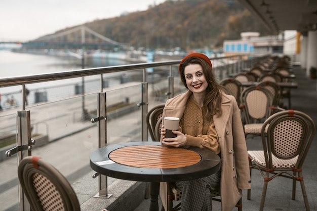 Feliz niña sentada al aire libre restaurante de la calle o terraza mirando sonriendo a otro lado sosteniendo un café ...