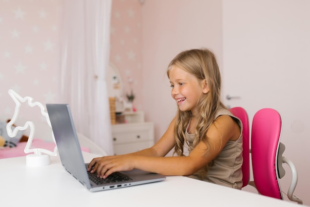 Una feliz niña de siete años está sentada en su portátil y se comunica con sus amigos a través de un enlace de video Aprendizaje a distancia en casa