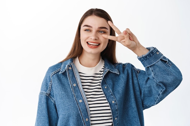 Feliz niña sincera, sonriendo y mirando a un lado el texto promocional, leyendo copyspace mientras muestra el signo v de paz, de pie positivo y optimista contra el fondo blanco