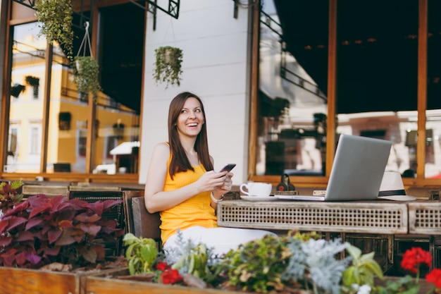 Feliz niña sonriente en la cafetería de la calle al aire libre sentado en la mesa con la computadora portátil, mensaje de texto en un amigo del teléfono móvil, en el restaurante durante el tiempo libre