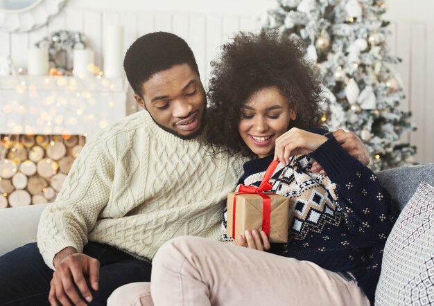 Feliz niña sonriente desenvolviendo el regalo de año nuevo de su novio cerca del árbol de navidad