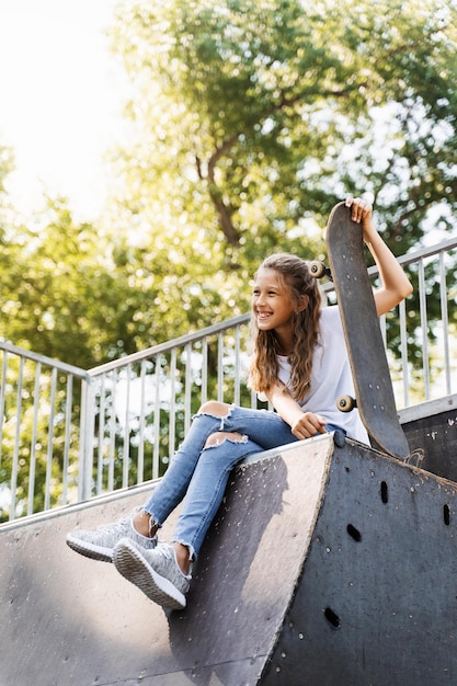 Feliz niña sonriente con patineta sentada en el patio de recreo y divirtiéndose Estilo de vida deportivo extremo Niño riendo con patineta posando en la rampa deportiva