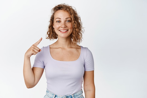 Feliz niña sonriente con pelo corto rubio, señalando con el dedo a sí misma, autopromoción, de pie en traje casual en blanco.