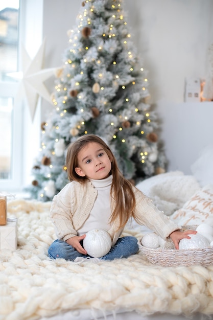 Feliz niña sonriente en suéter blanco con bolas de juguete de Navidad en la víspera de Navidad sentarse en la cama