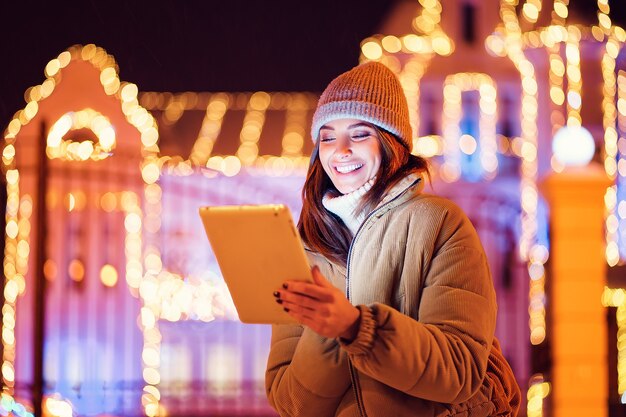 Feliz niña sonriente usa su tableta digital mientras está de pie al aire libre fondo de vacaciones de Navidad