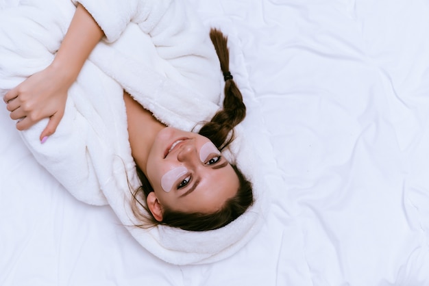 Feliz niña sonriente yace en una cama con una bata de felpa blanca