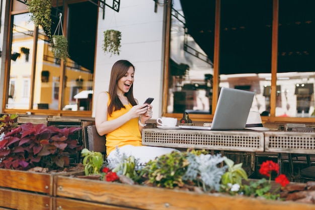 Feliz niña sorprendida en la cafetería de la calle al aire libre sentado en la mesa con la computadora portátil, mensaje de texto en un amigo del teléfono móvil, en el restaurante durante el tiempo libre