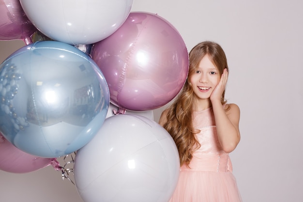 Feliz niña sorprendida con globos de aire pastel en estudio sobre fondo blanco.