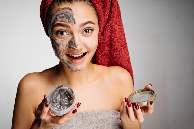 Feliz niña con una toalla roja en la cabeza se aplicó una útil mascarilla de arcilla a la mitad de la cara, se ríe