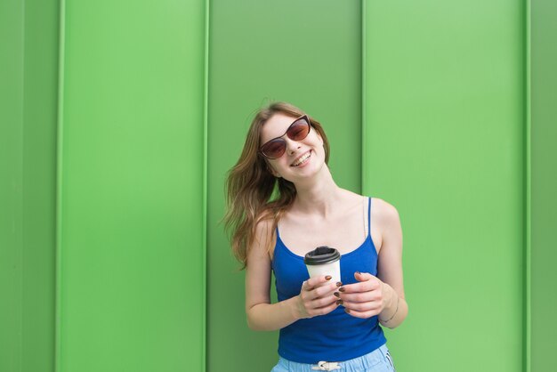 Feliz niña vestida con una camiseta azul se encuentra en el fondo de una pared verde con un vaso de café en sus manos, sonriendo y mirando a la cámara.