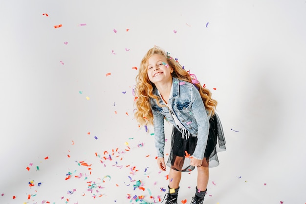 Feliz niña vestida a la moda con cabello rizado en chaqueta vaquera y falda tutú negra y botas ásperas en blanco con confeti de colores