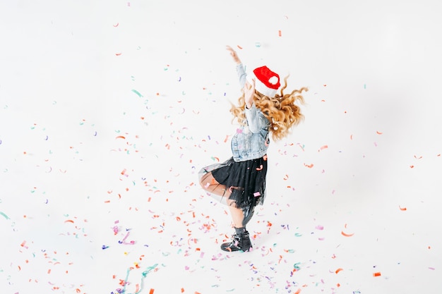 Feliz niña vestida a la moda con cabello rizado, sombrero de santa, chaqueta vaquera y falda tutú negra sobre blanco con confeti de colores