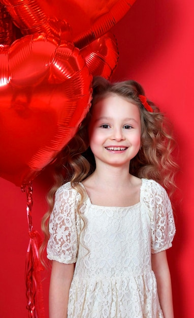 Feliz niño alegre positivo niña pequeño pequeño emocionado lindo niño en vestido blanco sosteniendo globos en forma de corazón rojo divirtiéndose y sonriendo sobre fondo rojo Presente Holliday Foto vertical