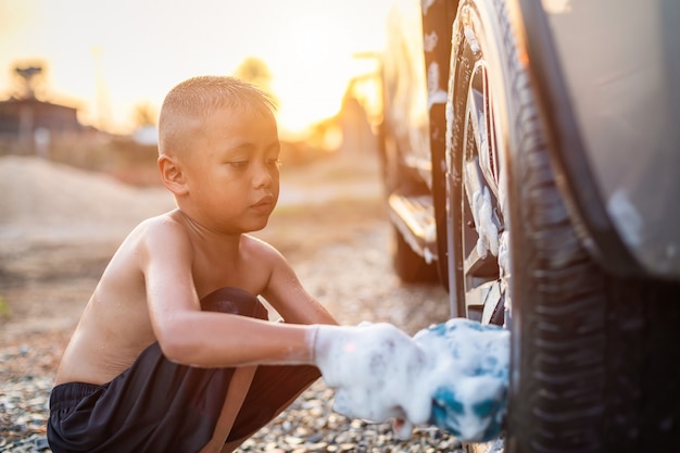 Feliz niño asiático con jabón blanco y el uso de una esponja azul para lavar el automóvil al aire libre en la hora del atardecer