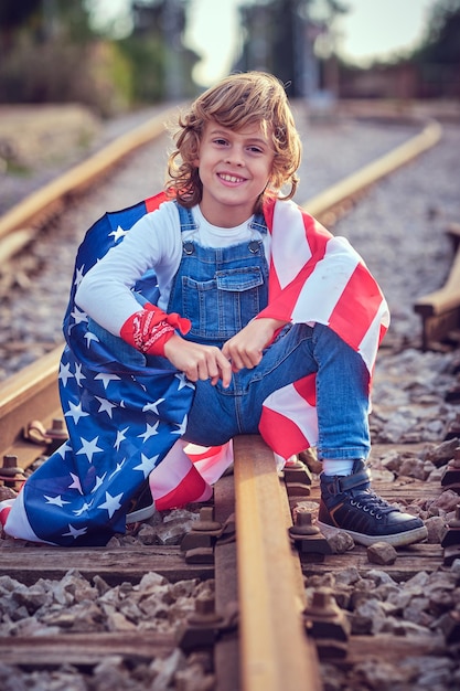 Feliz niño de cabello blanco con la bandera nacional de los Estados Unidos en tela de vaqueros mirando a la cámara mientras está sentado en el interruptor ferroviario