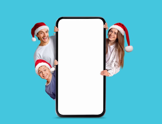 Feliz niño caucásico y pareja milenaria con sombreros de Santa Claus con un gran smartphone con pantalla vacía