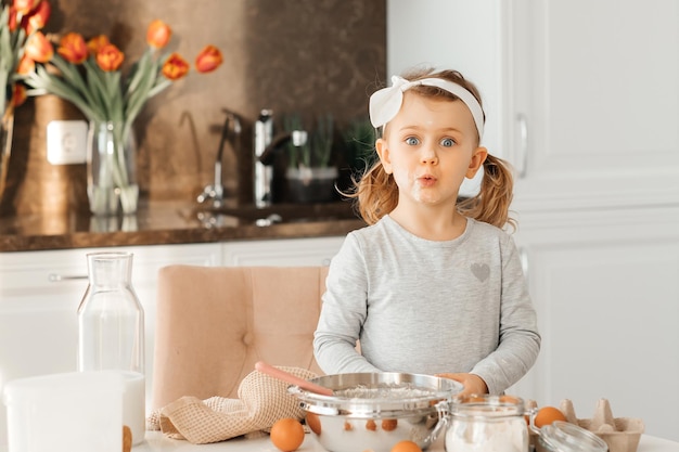 Feliz niño emocionado niña horneando pastel para Pascua niño emocional cocinar postre presente para el día de la madre en cocina blanca soleada Panadería casera Botella de leche huevos harina están sobre la mesa