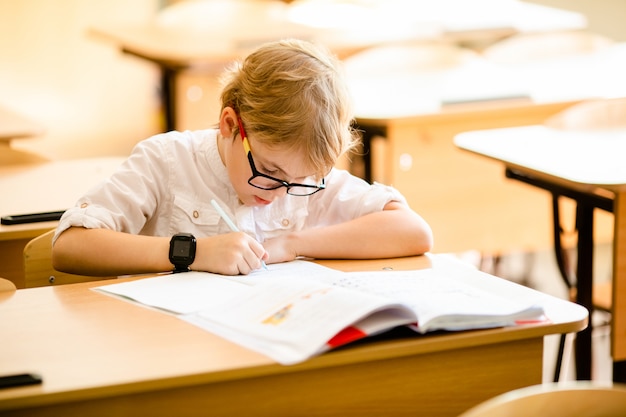 Feliz niño inteligente lindo está sentado en un escritorio en unas gafas con levantar la mano.