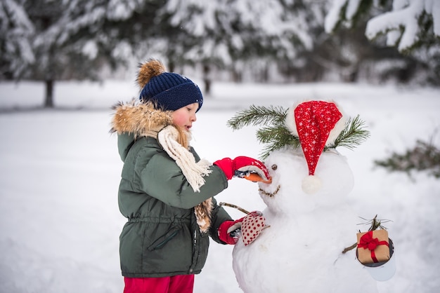 Feliz niño lindo rubio plaing con un muñeco de nieve en un paseo de invierno cubierto de nieve