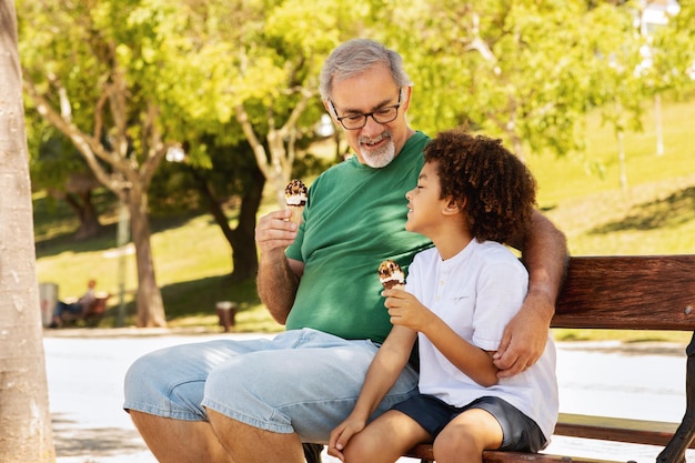 Feliz niño de raza mixta y viejo abuelo caucásico disfrutan de tiempo libre sentados en un banco comiendo hielo