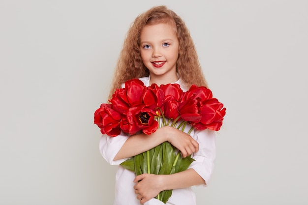 Feliz niño rubio con cabello ondulado mirando al frente con expresión feliz, sosteniendo tulipanes rojos, contento de obtener hermosas flores