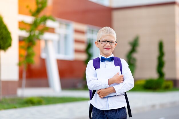 Feliz niño rubio colegial con gafas con una mochila se encuentra en la escuela y se ríe