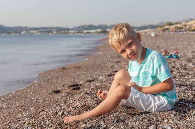 Feliz niño sonriente sentado en la playa y jugando con piedras