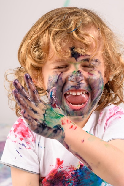 Feliz niño sucio cubierto de pinturas coloridas divirtiéndose mientras está sentado en un estudio ligero con los ojos cerrados sobre fondo blanco