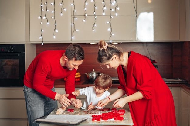 Feliz noite na véspera de Natal ou véspera de Ano Novo, a família de três pessoas está ocupada assando biscoitos, vestida de vermelho, cozinha aconchegante, atmosfera de amor e alegria. As pessoas se divertem.